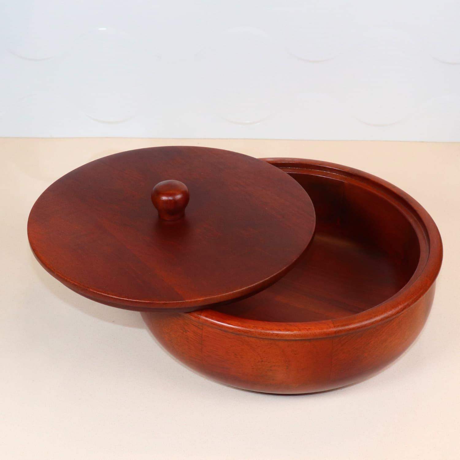 A round, polished wooden bowl with a matching lid partially open, both in a deep reddish-brown color, is placed on a light-colored surface. The bowl has a knob on the lid for easy handling. This premium The Woody Casserole Chapati box by LOOSEBUCKET stands out against the plain and subtle background.