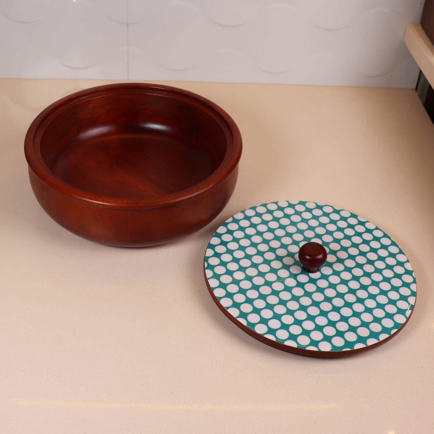 A polished wooden bowl, resembling *The Dot legacy Chapati Box* in India, features a separate round lid adorned with white polka dots on a teal background. The lid has a wooden knob handle, and both items rest elegantly on a smooth, light-colored surface.
