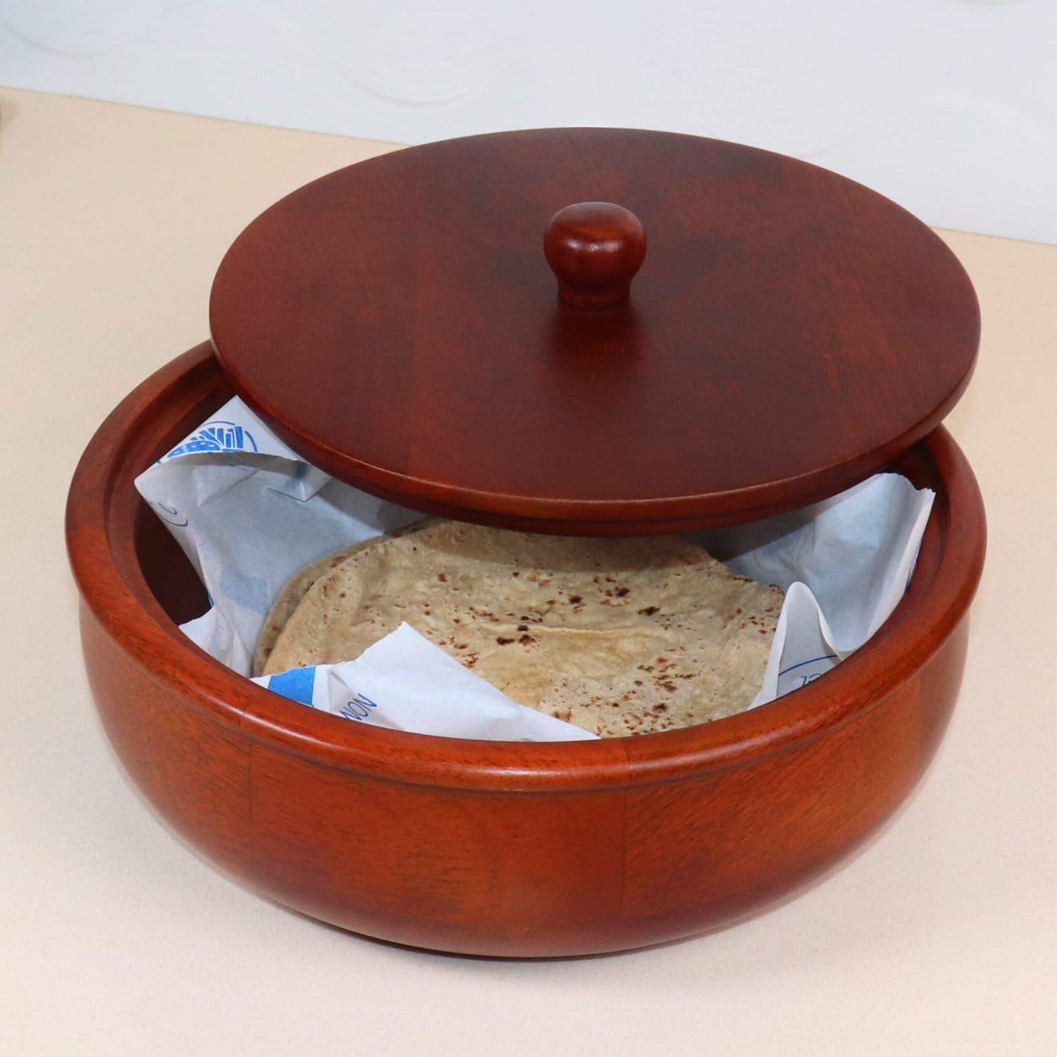 A round, wooden container, touted as the best *The Woody Casserole Chapati box* in India, with a lid partially open, shows a stack of tortillas inside wrapped in paper. The container is placed on a light-colored surface.