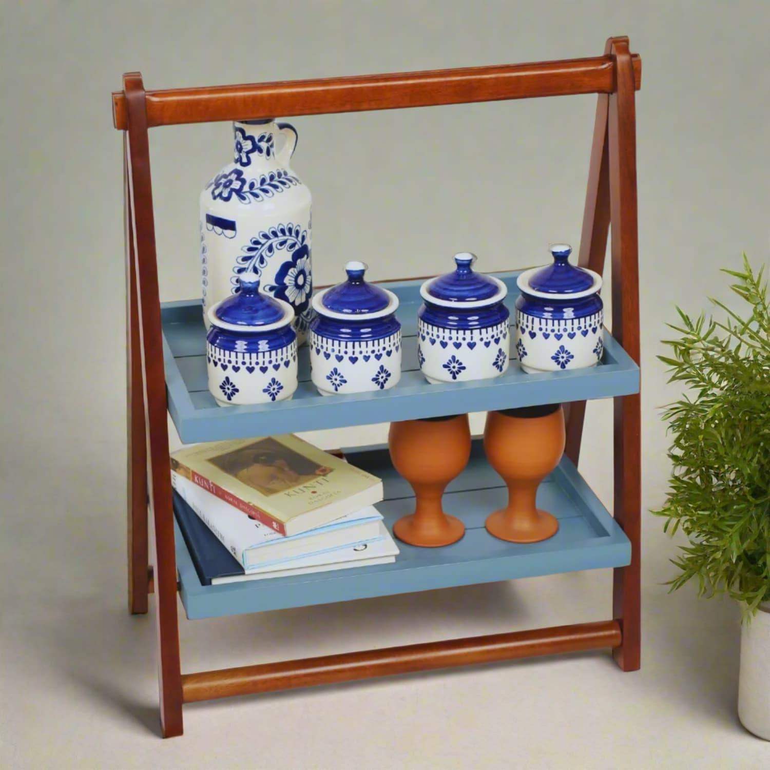 Loosebucket Foldable Kitchen Organizer displaying blue and white ceramic spice containers, terracotta cups, and books on two-tier mahogany frame with grey-blue shelves, styled with green plant decoration, perfect for kitchen storage and organization.